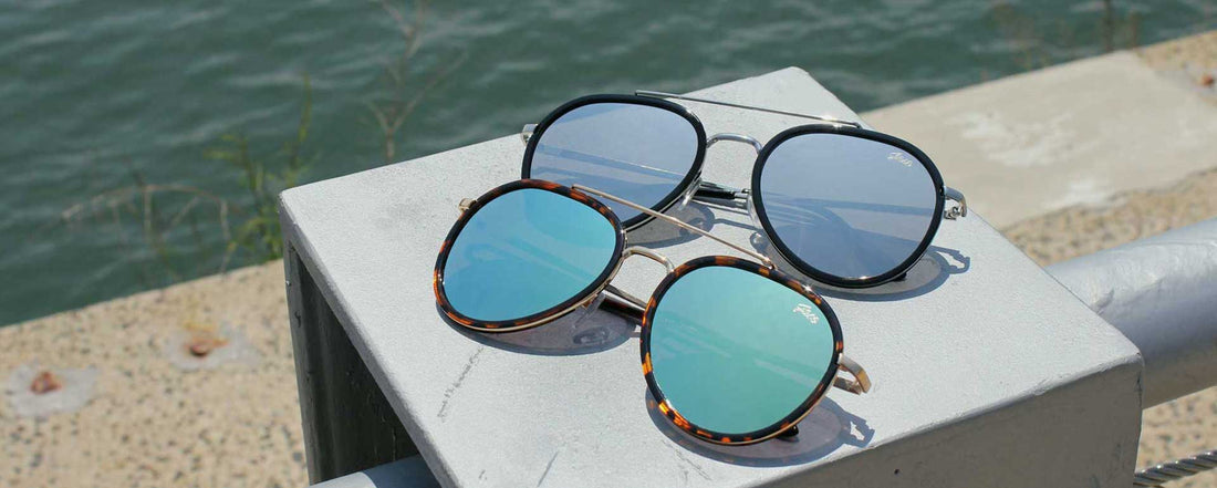 two pairs of aviator sunglasses with different color mirror lenses placed on a pillar near the waterfront