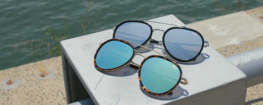 two pairs of aviator sunglasses with different color mirror lenses placed on a pillar near the waterfront