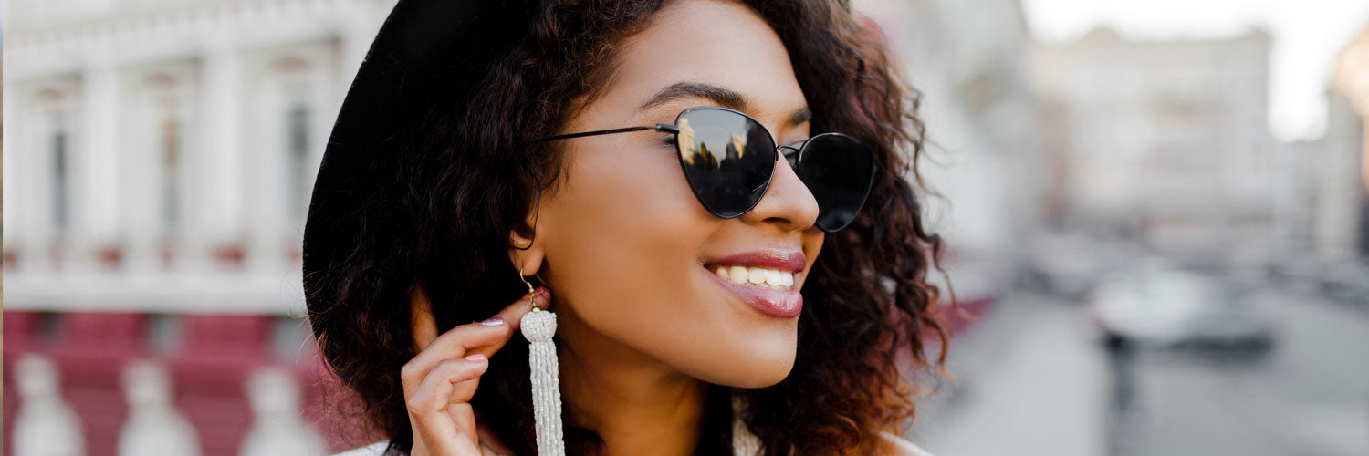 young black female wearing black cat eye sunglasses and hat looking to the right with white buildings in the background