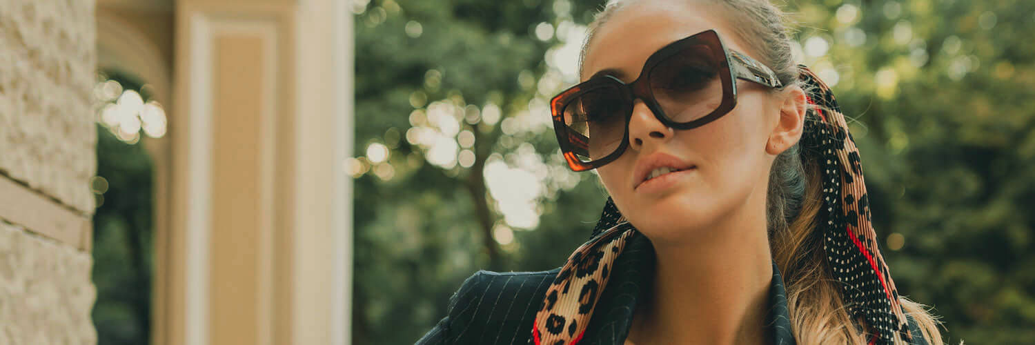 brunette woman wearing large tortoise shell sunglasses and striped blazer next to stone wall and trees