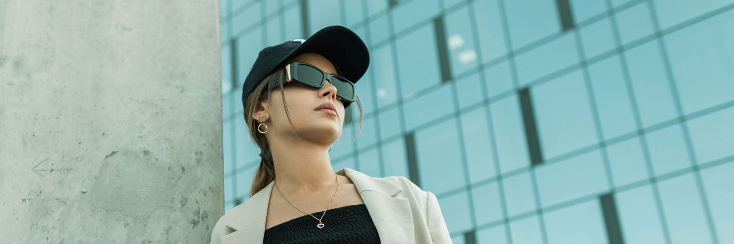 Brunette woman standing against concrete wall wearing black sunglasses and baseball cap looking to the right with a large all glass facade building in the background