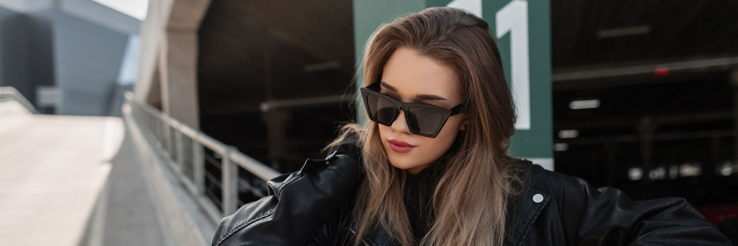 brunette woman wearing black sunglasses standing along a sloped pavement with building in the background