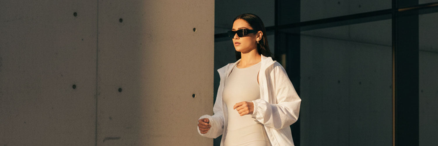 Woman in a white outfit standing against a concrete wall with sunlight casting shadows.